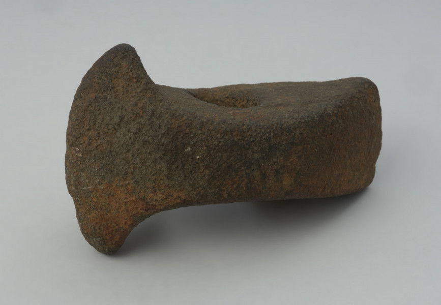 Topór kamienny - Ujęcie z przodu skosem w lewą stronę; Topór z szerokim, symetrycznie rozszerzonym ostrzem i nieznacznie rozszerzonym obuchem, o prostokątnym przekroju poprzecznym, wykonany z szarego surowca skalnego. Otwór niemal kolisty, cylindryczny. Powierzchnia silnie zerodowana. Drobne uszkodzenie ostrza.