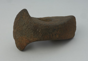 Topór kamienny - Ujęcie z przodu skosem w lewą stronę; Topór z szerokim, symetrycznie rozszerzonym ostrzem i nieznacznie rozszerzonym obuchem, o prostokątnym przekroju poprzecznym, wykonany z szarego surowca skalnego. Otwór niemal kolisty, cylindryczny. Powierzchnia silnie zerodowana. Drobne uszkodzenie ostrza.