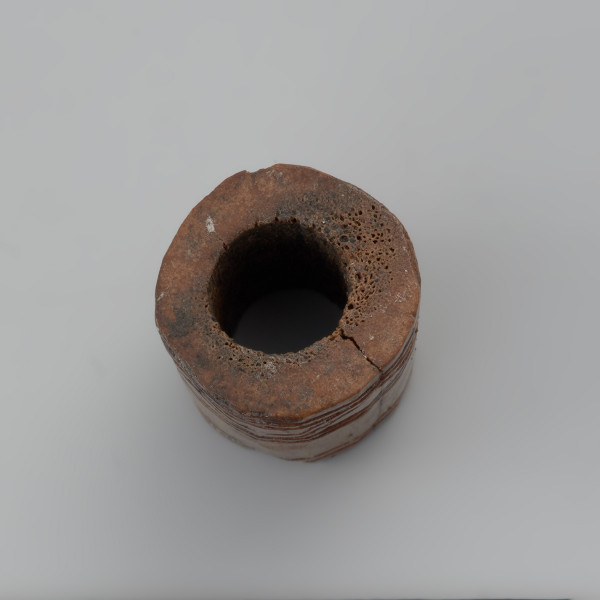 oprawka stożkowata - Ujęcie z góry oprawki. Oprawka ma stożkowaty kształt o ściętym wierzchołku. Ślady pozostawione na jej powierzchni świadczą o wykorzystaniu do jej produkcji co najmniej trzech narzędzi – piły, noża oraz świdra. Na powierzchni oprawki znajduje się mało skomplikowany wzór zdobniczy o motywie liniowym naniesionym za pomocą ostrego narzędzia. Górną część zajmuje ornament w postaci dwóch dookolnych linii rytych. Natomiast na przeciwległej stronie występuje ozdobnik w formie linii zygzakowatych wpisanych od góry i od dołu w podwójne linie ryte.