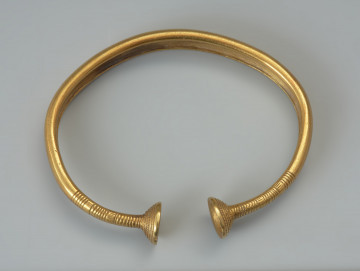 Bransoleta z pieczątkowatymi zakończeniami - Ujęcie z tyłu lekkim skosem w lewo. Bransoleta otwarta wykonana ze złotej, grubej blachy o brzegach zagiętych pod spód. Zwieńczona jest pieczątkowatymi zakończeniami, przypominającymi naparstki lub stożkowate miseczki. Zakończenia zdobione są dookolnym żebrowaniem, a brzegi kabłąka grupami nacięć, układających się w motyw metopowy.