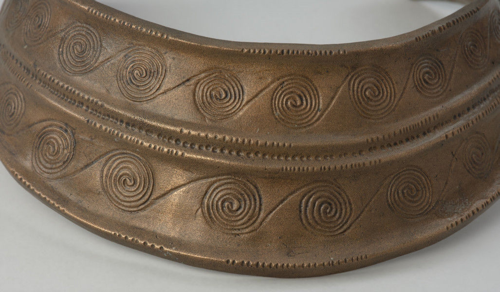 Brązowy naszyjnik kołnierzowaty - Ujęcie detalu przodu naszyjnika. Brązowy naszyjnik kołnierzowaty jest zdobiony motywem  spiralnych pasm ułożonych w dwóch rzędach, oddzielonych podwójnym żeberkiem, ornamentowanym
drobnymi nacięciami i punktowaniem.