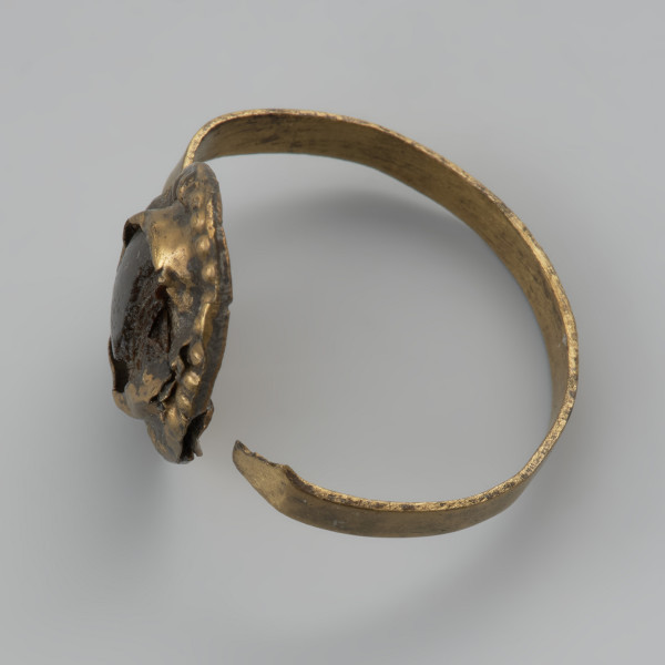 pierścionek z oczkiem - Ujęcie prawej strony z góry. Trzyczęściowy brązowy pierścionek z oczkiem, składający się z metalowej taśmy stanowiącej otok, metalowej tarczki pod oczko złożonej z owalnej płaskiej blaszki spodniej oraz dodatkowej taśmy tworzącej eliptyczne gniazdko. Jest ona zdobiona dookoła odciskami. Trzecim elementem, który uzupełnia oczko jest grafitowe szkiełko zachowane jedynie w części, przyklejone do spodniej tarczki i przytrzymywane przez krawędzie otaczającej je taśmy. Między blaszkami oczka pozostawione są dwie 