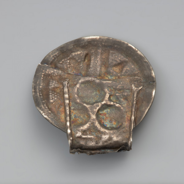 ozdoba; biżuteria; zawieszka - Ujęcie tyłu zawieszki uszkiem do dołu. Okrągła zawieszka wycięta ze srebrnej blachy o średnicy 2,5 cm, wraz z pasmem zawiniętym w rurkowate uszko. Zdobiona jest po jednej stronie trzema guzkami, z których każdy jest otoczony wieńcem przylutowanych półkolistych granulek. Również na każdym z guzków znajdują się po trzy granulki. Na pozostałej powierzchni blaszki znajdują się cztery promieniście rozłożone pola wypełnione ornamentem trójkątów wykonanym w technice granulacji oraz łańcuszkiem półjodełki. Uszko, również zdobione jest granulkami. Do tylnej, niezdobionej strony, od boków uszka do około połowy wysokości okrągłej części, przylutowane są wąskie pasemka blachy wzmacniające zawieszkę. Zarówno uszko jak i zawieszka właściwa poniżej linii guzków są pęknięte.