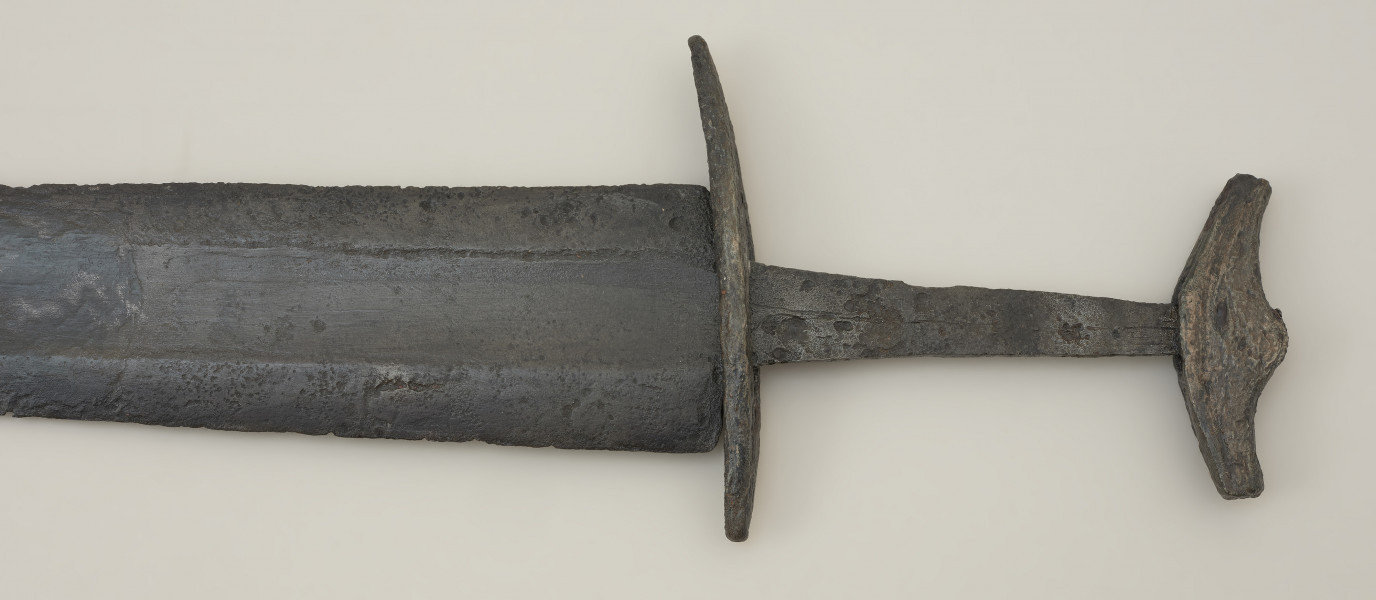 miecz dwusieczny - Ujęcie rękojeści miecza z przodu. Całkowicie zachowany, z niewielkimi ubytkami korozyjnymi w okolicach sztycha. Jego głownia jest najszersza przy jelcu i równomiernie zwężą się ku ostremu sztychowi. Jest zaopatrzona w szerokie zbrocze. Głownię wykonano w technice prętowej, poprzez zgrzanie stalowych nakładek ostrzowych z jednolitym rdzeniem. Trzpień mocno zwęża się ku głowicy. Jelec, soczewkowaty w rzucie z góry, jest długi i płaski, a jego końce są zakrzywione w kierunku głowni. Jednolita głowica ma kształt siodełkowaty. Jej dolna część wygięta jest ku górze. Na jej powierzchni zaznaczono symboliczny podział na podstawę i nakładkę.