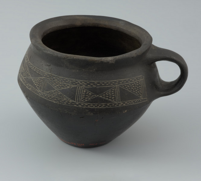 Naczynie gliniane - Ujęcie lewego boku. Naczynie gliniane (kubek) posiada gładką, czarną powierzchnię i drobną domieszkę.