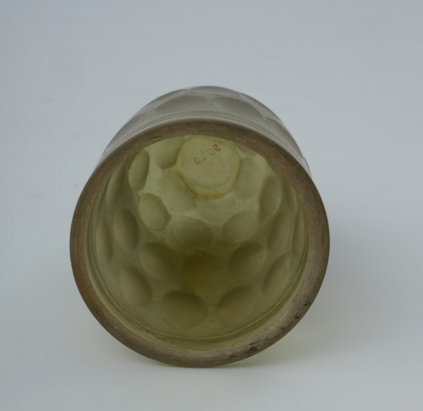 Pucharek szklany barwy jasnozielonej - Ujęcie z góry. Pucharek szklany barwy jasnozielonej. Naczynie jest grubościenne zdobione czterema rzędami szlifowanych owali obiegających pucharek dookoła oraz dwoma żłobieniami pod krawędzią.