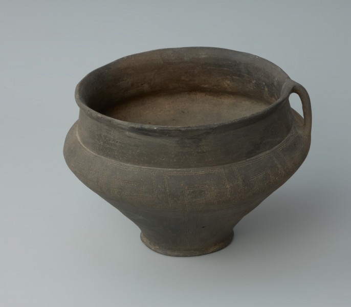 Ujęcie z przodu. Waza gliniana z uchem o podciętej dolnej partii brzuśca i wysokim załomie. Szyja naczynia jest niemal cylindryczna, wylew delikatnie wychylony na zewnątrz i zaokrąglony. Waza jest ciebnobrunatna, o wygładzanej powierzchni. Naczynie jest zdobiona dookolnym pasem ornamentacyjnym w postaci powielonych swastyk wykonanym kółkiem zębatym.