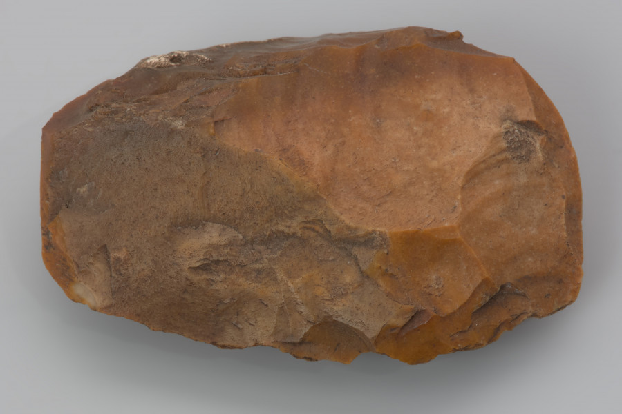 ciosak odłupkowy - ujęcie z góry; Ciosak wykonany z odłupka krzemienia kredowego. Na obu bokach ciosaka zachowały się fragmenty oryginalnej strony wierzchniej i spodniej odłupka, z którego został wykonany. Przekrój soczewkowaty. Na bokach widoczne negatywy odbić kształtujących wykonanych miękkim tłukiem. Ostrze uformowane 3 odbiciami. Zabytek pokryty jest pomarańczowo-czerwoną patyną będącą efektem depozycji w środowisku o wysokiej zawartości żelaza.