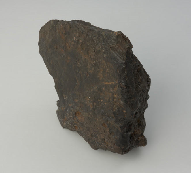 bryła metalu - Ujęcie w pionie tyłu. Fragment placka odlewniczego o nieregularnym, czworobocznym kształcie z zachowanym jednym z pierwotnych boków o ostrej krawędzi. W przekroju poprzecznym trójkatny.