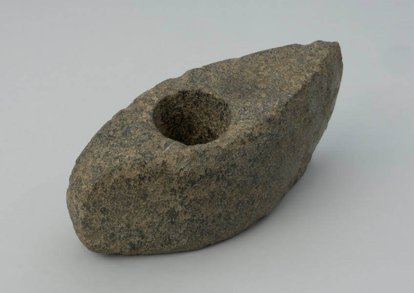 Topór kamienny - Ujęcie z góry skosem w prawą stronę. Topór o wydłużonej części tylnej zakończonej wąskim obuchem, z wywierconych otworem przesuniętym ku tyłowi. Przekrój poprzeczny czworokątny wysoki. W w rzucie bocznym trapezowaty z lekko skośnym ostrzem i lekko łukowatym obuchem. Przy otworze, na stronie górnej, ślady wykucia płytkiej miseczki pod wiercenie. Powierzchnia zerodowana ze śladami szlifowania i puncowania. Ostrze wtórnie uszkodzone