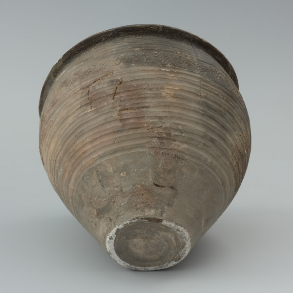 naczynie gliniane - Ujęcie spodu w poziomie. Garnek o baniastej formie z charakterystycznie wychyloną na zewnątrz krawędzią wylewu, obtoczony w całości na kole garncarskim. Brzusiec garnka znajduje się w 2/3 wysokości. Powierzchnia glinianego pojemnika przyozdobiona jest ornamentem wielowątkowym skośnych żłobków, dookolnych linii rytych, linii falistych oraz ponownych linii rytych. Fakturę naczynia tworzy dobrze wyprofilowana, gładka powierzchnia. Garnek wylepiono z glinianych wałeczków z wcześniej przygotowanej masy garncarskiej zawierającej domieszkę drobno i średnioziarnistego tłucznia - piasku i granitu.