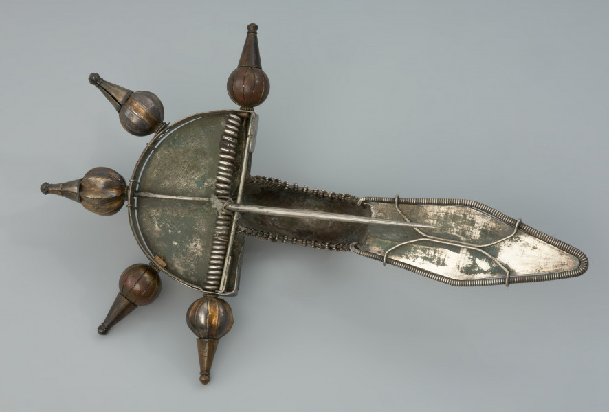 ozdoba; Srebrna, pozłacana zapinka - Ujęcie z dołu. Srebrna, pozłacana fibula z rombowato ukształtowaną długą nóżką, krótką pochewką i wysoko wysklepionym kabłąkiem. Zapinka ma główkę w postaci półkolistej tarczki nad sprężynką zaopatrzoną w 5 guzów wykonanych ze złotej taśmy.