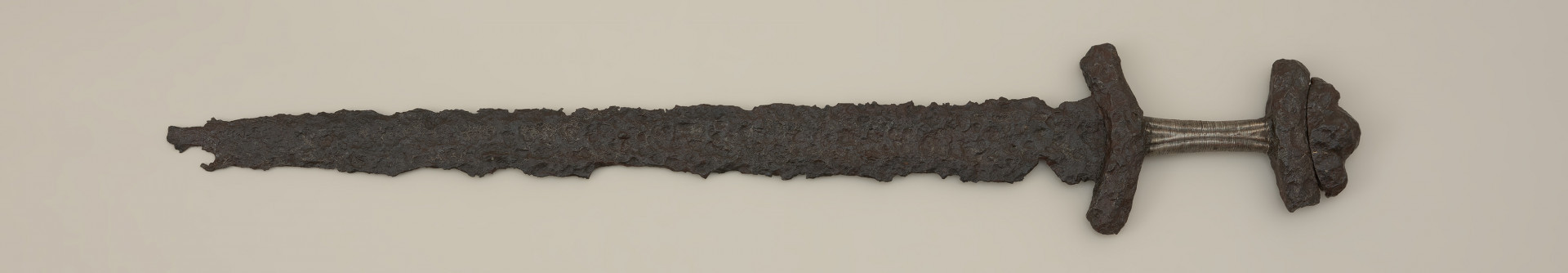 miecz dwusieczny - Ujęcie z przodu. Dwusieczna, mocno skorodowana głownia zachowała się w około 2/3 pierwotnej długości z czytelnym szerokim zbroczem. Najszersza jest w okolicach zastawy i równomiernie zwęża się ku sztychowi. Elementy rękojeści wykonano z żelaza. Jelec, łódkowaty w rzucie z góry, ma kształt sztabkowy o ramionach wygiętych w kierunku głowni. Ramiona podstawy głowicy zwrócone są ku górze. Na nich osadzono nakładkę o trójdzielnej formie. Jej część środkowa jest większa od pozostałych. Trzpień okręcono srebrnym drutem, tworzącym tzw. ornament schodkowy po jednej stronie i znak X po drugiej.