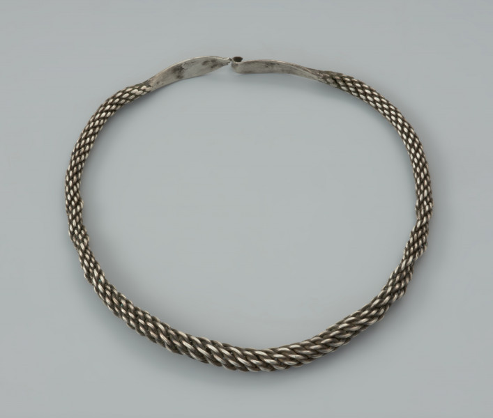 naszyjnik srebrny - Ujęcie z góry przodu zapiętego naszyjnika. Srebrny naszyjnik spleciony z pięciu par drutów owiniętych wokół siebie, z końcami rozklepanymi w owalne, niezdobione płytki. Jedna z nich zakończona została haczykiem, końcówka drugiej jest ułamana. W części środkowej druty są skręcone luźniej, co powoduje, ze obręcz jest w tym miejscu grubsza, natomiast po obu stronach znajdują się fragmenty, gdzie splot jest nierówny.