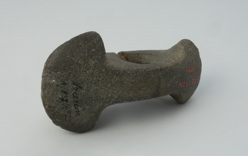 broń obuchowa - Ujęcie z boku. Topór kamienny obosieczny, tzw. o dwóch ostrzach, labrys lub topór Amazonek, na obu końcach rozszerzony symetrycznie w górę i dół, przy czym koniec ostrzejszy jest słabiej rozchylony, niż druga strona. Otwór do osadzenia trzonka znajduje się około połowy długości korpusu. Przełamany na wysokości otworu i sklejony. Powierzchnia gładzona, nieco zniszczona