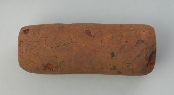 zawieszka cylindryczna; amulet bursztynowy - Ujęcie w poziomie przodu zawieszki. Podłużna, cylindryczna zawieszka wykonana z dużej konkrecji bursztynu, której większość powierzchni pokrywa patyna przypominająca glinę. W szczytowej oraz bocznej ściance znajdują się wywiercone otworki nie przechodzące przez całą grubość przedmiotu.