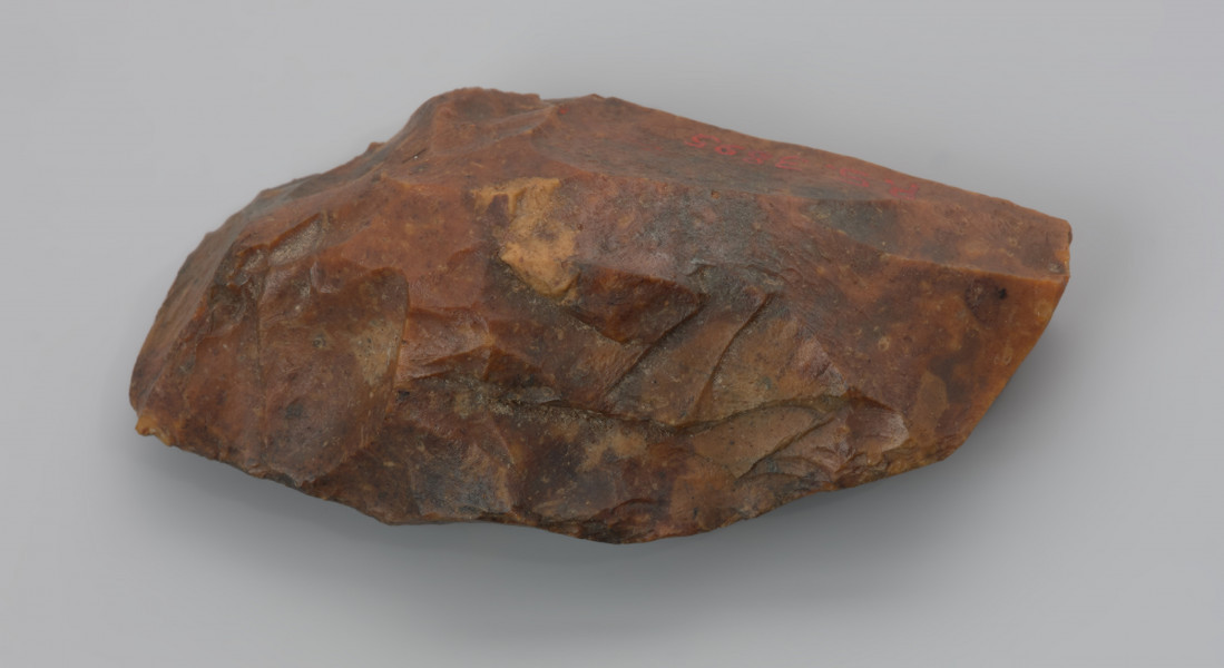 Mały ciosak rdzeniowy - ujęcie z boku; Na zdjęciu ciosak rdzeniowy, narzędzie wykonano z krzemienia kredowego. Pokryte jest czerwonopomarańczową patyną, która jest efektem zalegania w środowisku bogatym w związki żelaza.