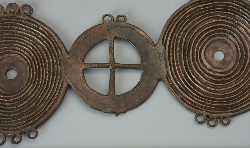 Zapinka trójelementowa - Ujęcie fragmentu zapinki. Zapinka składa się z dwóch zewnętrznych i jednej centralnej tarczki. Dwie zewętrzne pokrywają koncentryczne żłobki. Na jednej z nich pośrodku znajduje się ozdobny detal w formie guzka. Analogiczny guzek na drugiej tarczce nie zachował się. Na trzeciej, wewnętrznej tarczce o kolistym kształcie wpisany jest krzyż. Na stronie spodniej znajduje się igła będąca elementem zapięcia.