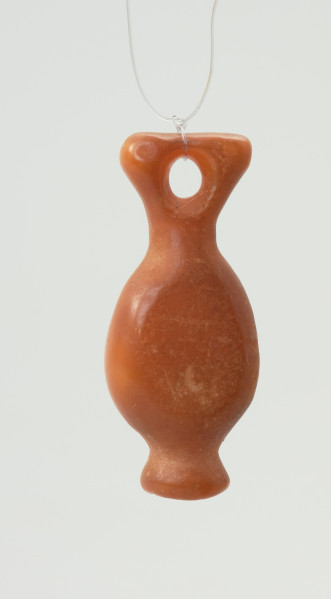 zawieszka bursztynowa - Ujęcie z tyłu w pionie. Zawieszka z bursztynu o pomarańczowej mlecznej barwie ma kształt spłaszczonej po bokach ryby z dziurką w miejscu płetwy ogonowej, o całkowitej długości 3,3 cm. Powierzchnie są wygładzone, z niewielkimi uszkodzeniami oraz ubytkiem od strony głowy.