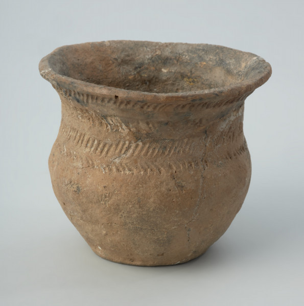 Naczynie gliniane - Ujęcie z przodu; Przysadzisty puchar o szerokim wylewie, zdobiony na szyjce czterema dookolnymi rzędami ukośnych, podłużnych odcisków, które tworzą pasma złożone z dwóch szeregów odcisków układających się w motyw rybich ości. Ukośnymi nacięciami jest zdobiona także krawędź wylewu.