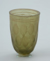 Pucharek szklany barwy jasnozielonej - Ujęcie z przodu. Pucharek szklany barwy jasnozielonej. Naczynie jest grubościenne zdobione czterema rzędami szlifowanych owali obiegających pucharek dookoła oraz dwoma żłobieniami pod krawędzią.