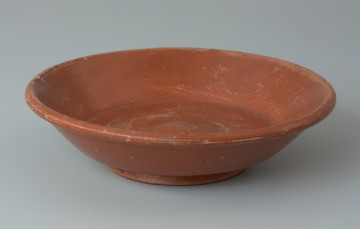 Rzymski talerz z gliny wykonany w technice terra sigillata - Ujęcie z przodu z góry. Rzymski talerz z gliny wykonany w technice terra sigillata. Naczynie jest gładkie, barwy ceglastej.