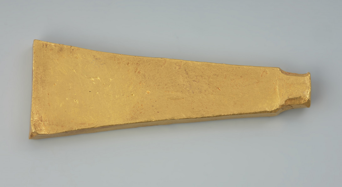 Sztabkowata płytka ze złota - Ujęcie z tyłu. Sztabkowata płytka ze złota w formie symetrycznego klina przechodzącego u szczytu w imitację owalnej w przekroju nasady lub ucha. Płytka jest zdobiona tylko na stronie wierzchniej.