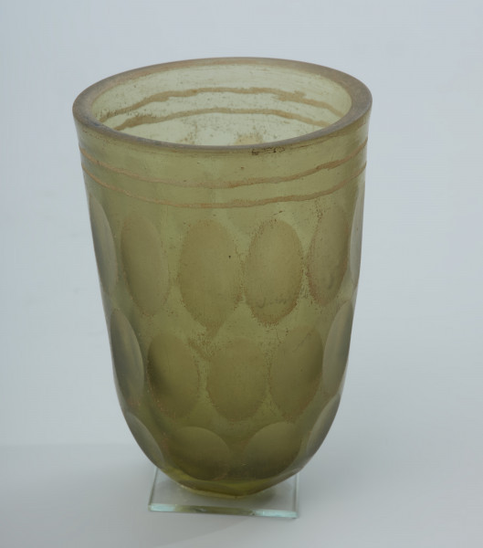 Pucharek szklany barwy jasnozielonej - Ujęcie z boku. Pucharek szklany barwy jasnozielonej. Naczynie jest grubościenne zdobione czterema rzędami szlifowanych owali obiegających pucharek dookoła oraz dwoma żłobieniami pod krawędzią.