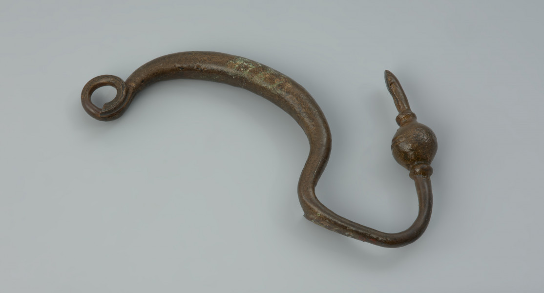 Kabłąk zapinki z brązu - Ujęcie z góry z tyłu. Zapinka z brązu; zachował się tylko kabłąk jednoczęściowy, przechodzący w odgiętą w górę nóżkę, zachowany jest jeden zwój sprężynki.