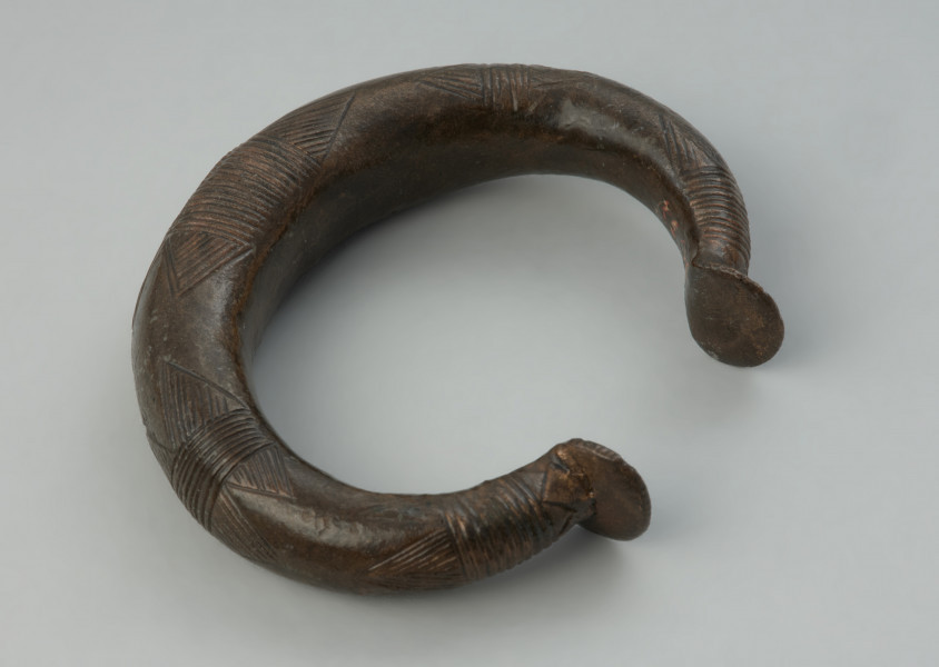 Bransoleta obręczowa - Ujęcie z góry prawej strony bransolety. Masywna bransoleta obręczowa o owalnym kształcie, odlana z brązu w formie grubego pręta. Jej zakończenia mają kształt niewielkich stopek. Powierzchnię obręczy zdobią grupy poprzecznych kresek oraz motyw tzw. wilczych zębów.