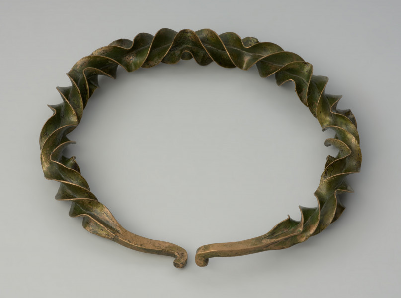 naszyjnik wieńcowaty typu Wendelring - Ujęcie z góry tył.u naszyjnika Naszyjnik formą przypominający wieniec wykonano z brązowego, silnie rozklepanego na krawędziach, czworograniastego pręta o gwiaździstym przekroju, który skręcono (tordowano) wokół własnej osi zmieniając kilkukrotnie kierunek skrętu, uzyskując efekt wąskich, pofałdowanych płaszczyzn o ostrych krawędziach wzdłuż niemal całej długości obręczy. Końce ukształtowane zostały w haczyki służące zapięciu.