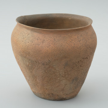 naczynie gliniane - Ujęcie z przodu. Garnek o jajowatym kształcie z wychylonym na zewnątrz wylewem, umocowanym na krótkiej szyjce. Największa wydętość brzuśca wypada w 2/3 wysokości garnka. W części przydennej od strony wewnętrznej zachowały się ślady produkcyjne w postaci pojedynczych dołków palcowych powstałych w wyniku formowania ścianek. Powierzchnia garnka jest nierówna i szorstka w dotyku, bez zdobień. Naczynie wylepiono ręcznie bez użycia koła garncarskiego, najprawdopodobniej z szerokich taśm odpowiednio przygotowanej gliny, wzbogaconej między innymi domieszką schudzającą średnioziarnistego piasku i granitu.