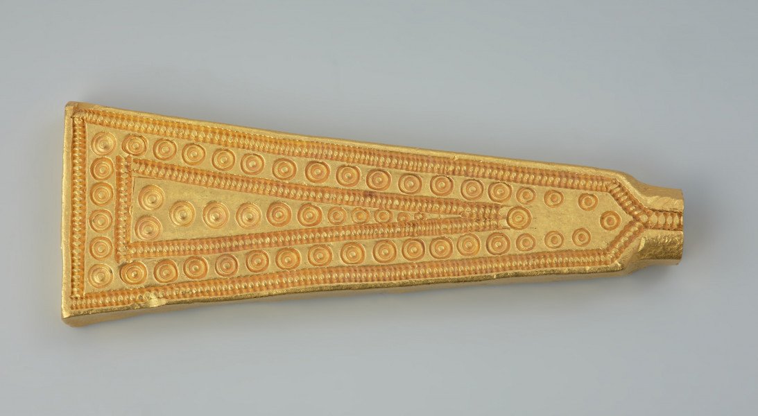 Sztabkowata płytka ze złota - Ujęcie z przodu. Sztabkowata płytka ze złota w formie symetrycznego klina przechodzącego u szczytu w imitację owalnej w przekroju nasady lub ucha. Płytka jest zdobiona tylko na stronie wierzchniej.