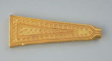 Sztabkowata płytka ze złota - Ujęcie z przodu. Sztabkowata płytka ze złota w formie symetrycznego klina przechodzącego u szczytu w imitację owalnej w przekroju nasady lub ucha. Płytka jest zdobiona tylko na stronie wierzchniej.