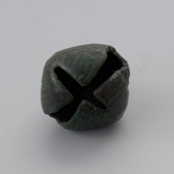 zawieszka dzwoneczkowata z Wolina - Ujęcie spodu zawieszki. Zawieszka dzwoneczkowata wykonana z brązowej blachy, spatynowanej na ziolono. Korpus przypomina kształtem pustą w środku kroplę rozciętą u dołu na krzyż z jednym ramieniem dłuższym i szerszym od drugiego. Dodatkowo dolna powierzchnia jest zdobiona żłobkowaniem. W rzucie z góry zawieszka ma kształt zbliżony do rąbu, u góry posiada otwór z trójkątnego w przekroju drutu. W jej wnętrzu znajduje się brązowa kulka. Powierzchnia dzwoneczka jest lekko zniszczona, a w ściance znajduje się niewielki ubytek.