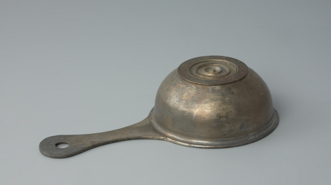 naczynie; Rzymski brązowy rondelek - Ujęcie z dołu z lewego boku. Rzymski brązowy rondelek z płaskim dnem o przewężonym uchwycie z otworem. Rondel jest zdobiony pod wylewem płytkimi dookolnymi liniami rytymi.