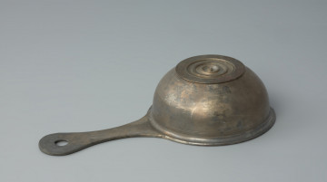 naczynie; Rzymski brązowy rondelek - Ujęcie z dołu z lewego boku. Rzymski brązowy rondelek z płaskim dnem o przewężonym uchwycie z otworem. Rondel jest zdobiony pod wylewem płytkimi dookolnymi liniami rytymi.