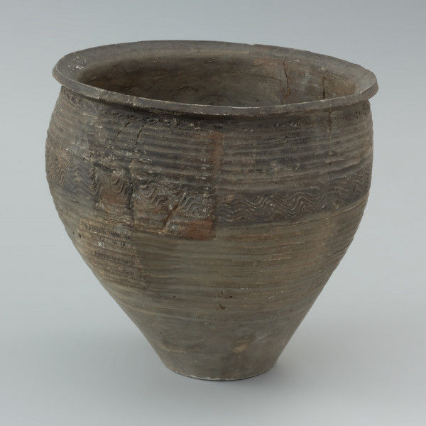 naczynie gliniane - Ujęcie lewej strony. Garnek o baniastej formie z charakterystycznie wychyloną na zewnątrz krawędzią wylewu, obtoczony w całości na kole garncarskim. Brzusiec garnka znajduje się w 2/3 wysokości. Powierzchnia glinianego pojemnika przyozdobiona jest ornamentem wielowątkowym skośnych żłobków, dookolnych linii rytych, linii falistych oraz ponownych linii rytych. Fakturę naczynia tworzy dobrze wyprofilowana, gładka powierzchnia. Garnek wylepiono z glinianych wałeczków z wcześniej przygotowanej masy garncarskiej zawierającej domieszkę drobno i średnioziarnistego tłucznia - piasku i granitu.