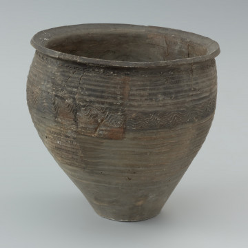 naczynie gliniane - Ujęcie lewej strony. Garnek o baniastej formie z charakterystycznie wychyloną na zewnątrz krawędzią wylewu, obtoczony w całości na kole garncarskim. Brzusiec garnka znajduje się w 2/3 wysokości. Powierzchnia glinianego pojemnika przyozdobiona jest ornamentem wielowątkowym skośnych żłobków, dookolnych linii rytych, linii falistych oraz ponownych linii rytych. Fakturę naczynia tworzy dobrze wyprofilowana, gładka powierzchnia. Garnek wylepiono z glinianych wałeczków z wcześniej przygotowanej masy garncarskiej zawierającej domieszkę drobno i średnioziarnistego tłucznia - piasku i granitu.