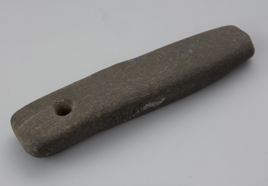 osełka kamienna - Ujęcie z góry. Osełka jest obłego kształt o owalnym zarysie z zaokrąglonymi krawędziami. Na jednym z końców znajduje się otwór przelotowy służący do zawieszania przedmiotu do paska albo na szyi. Na dolnej, górnej oraz bocznej powierzchni można zaobserwować ślady użytkowania podczas ostrzenia.