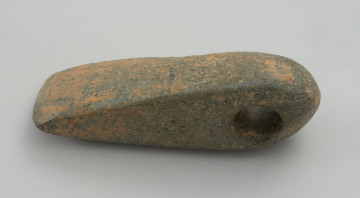 topór kamienny - Ujęcie lewego boku; Topór kamienny w typie pięciobocznych, ale z częścią obuchową bardziej zaokrągloną niż załamaną, z otworem umieszczonym w części obuchowej. W rzucie bocznym trapezowaty, z korpusem poszerzającym się dołem w stronę ostrza. Przekrój poprzeczny czworościenny o zaokrąglonych narożnikach. Otwór wiercony dwustronnie z widoczną poprawą kierunku wiercenia od dołu. Powierzchnia szlifowana, miejscami do połysku. Na stronie dolnej widoczne niedogładzenia.