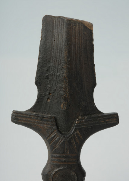 broń, miecz - Ujęcie detalu głowicy z przodu. Brązowy miecz z czworokatną, dekorowaną żłobieniami rękojeścią, głowicą w formie owalnej tarczki oraz jelcem ze skrzydełkami i półkolistym wycięciem.