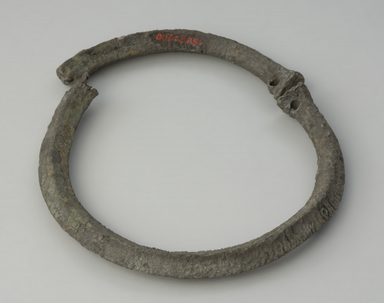 obręcz z czworograniastej sztaby - Ujecie obręczy z góry skosem z przodu. Srebrzystoszara obręcz, rozłamana na dwie części, wykonana została z czworograniastej cynowej sztaby. Przypomina kształtem naszyjnik obręczowy z ściętymi prosto i nieco pogrubionymi końcami oraz niewielkimi, nieregularnie rozmieszczonymi otworami. Zabytek jest zdeformowany, a jego powierzchnia popękana, co może świadczyć o wtórnym przepaleniu w ogniu.