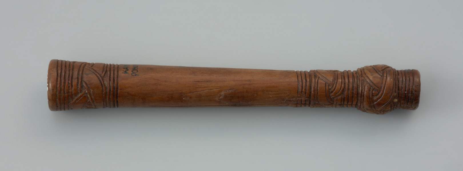 oprawka drewniana - Ujęcie z góry prawej strony oprawki. Drewniany trzonek podłużnego kształtu zbliżony do formy cylindrycznej. Na jednym z końców znajduje się kwadratowy otwór. Na obu przeciwległych stronach występują zdobienia w postaci prostej plecionki (warkocza) oraz dookolnych linii rytych. Zarówno trzonek jak i sam ornament wykonano z wielką starannością używając do tego ostrego noża.