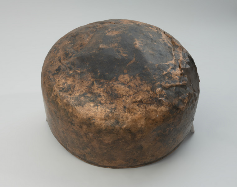 Kocioł ze stopu miedzi - Ujęcie spodu. Kocioł ze stopu miedzi o owalnym kształcie z dwoma trójkątnymi uchami. Kocioł wykonano ze stopu miedzi. Jego kształt jest asymetryczny. W części górnej jest węższy i owalny, natomiast w części dolnej szerszy o zaokrąglonym dnie.