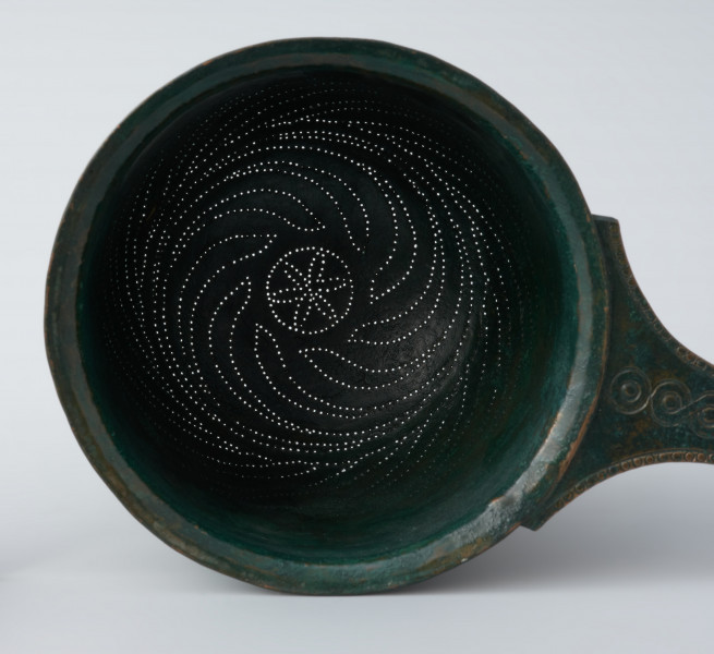 Ujęcie z góry w przybliżeniu. Rzymskie cedzidło z brązu z uchwytem zaopatrzonym w woluty.