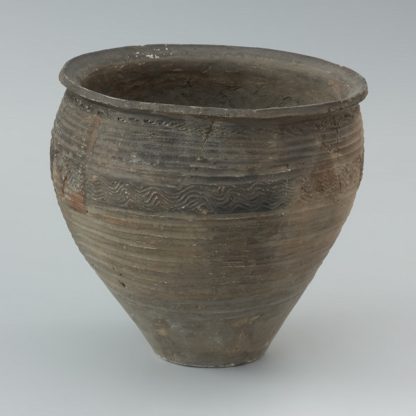 naczynie gliniane - Ujęcie prawej strony naczynia. Garnek o baniastej formie z charakterystycznie wychyloną na zewnątrz krawędzią wylewu, obtoczony w całości na kole garncarskim. Brzusiec garnka znajduje się w 2/3 wysokości. Powierzchnia glinianego pojemnika przyozdobiona jest ornamentem wielowątkowym skośnych żłobków, dookolnych linii rytych, linii falistych oraz ponownych linii rytych. Fakturę naczynia tworzy dobrze wyprofilowana, gładka powierzchnia. Garnek wylepiono z glinianych wałeczków z wcześniej przygotowanej masy garncarskiej zawierającej domieszkę drobno i średnioziarnistego tłucznia - piasku i granitu.