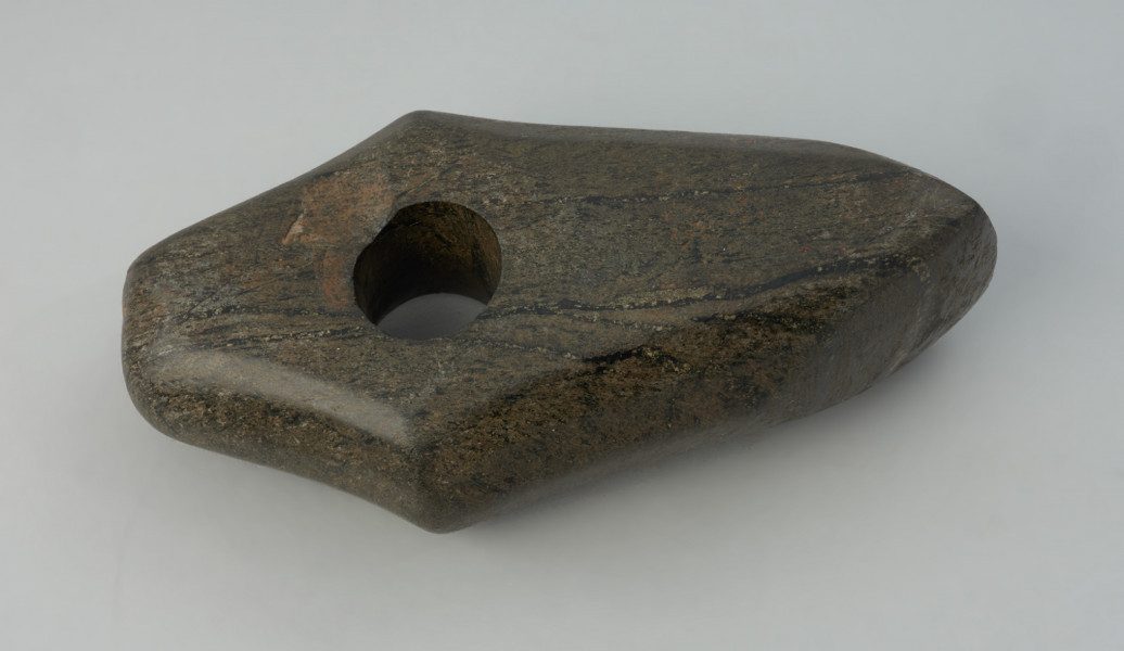 Topór kamienny - Ujęcie z góry.; Topór kamienny z wielopłaszczyznową, fasetowaną powierzchnią. Zarys pięcioboczny, ze zwężoną częścią tylną zakończoną tępym obuchem oraz ze „skrzydełkami” na wysokości przesuniętego nieco ku tyłowi otworu. Otwór stożkowaty, wiercony od jednej strony. Powierzchnia gładzona i polerowana. Ostrze ze śladami ponawianego ostrzenia, prawdopodobnie pierwotnie opuszczane (tzw. broda).