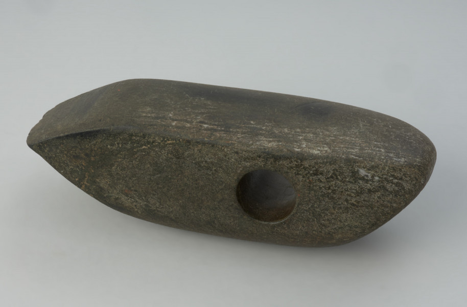 Topór kamienny - Ujęcie z boku ostrzem do tyłu; Asymetryczne narzędzie ze skośnym obuchem i otworem przewierconym równolegle do kierunku ostrza. W rzucie bocznym korpus jest prosty, przy czym część tylna jest nieco wyższa od przedniej. Otwór do osadzenia trzonka umieszczony bliżej części tylnej, jest wywiercony jednostronnie. W partii środkowej, opodal otworu, jest widoczne zaczątkowe wiercenie wykonywane okrągłym, pustym narzędziem, prawdopodobnie kością. Powierzchnia narzędzia jest gładzona w nierównym stopniu, przy otworze zauważalne są zagłębienia po odbiciach kształtujących ogólną formę narzędzia. W wielu miejscach zaobserwować można grupy równoległych rys, które są śladami po zdzieraniu i gładzeniu