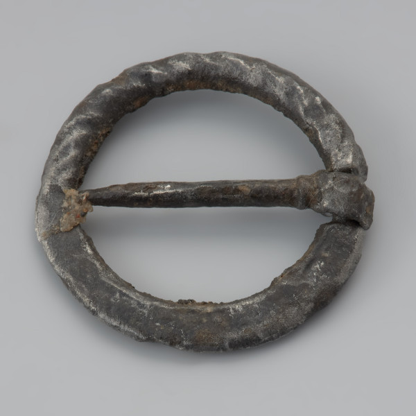 Sprzączka żelazna - Ujęcie z przodu z góry w lewą stronę. Sprzączka kolista, żelazna, z płaskim kolcem umieszczonym na wypłaszczeniu ramki. Jedna część ramki jest tordowana